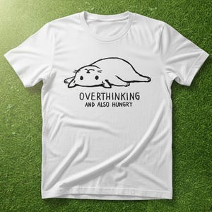 May include: White t-shirt featuring a black illustration of a seal-like animal. The text "OVERTHINKING AND ALSO HUNGRY" is printed below the image. The shirt is made of a soft material.