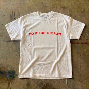 Puede incluir: Camiseta blanca con el texto rojo "DO IT FOR THE PLOT". La camiseta está hecha de un material suave, probablemente algodón. La camiseta está tendida sobre una superficie texturizada.
