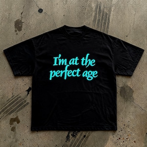 May include: A black short-sleeved t-shirt featuring the text "I'm at the perfect age" printed in a bright cyan blue script font across the centre of the chest. The garment has a crew neckline and a standard fit with short sleeves.
