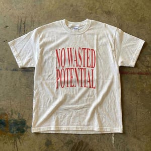 May include: A white t-shirt with the red text "NO WASTED POTENTIAL" printed on the front. The shirt has short sleeves and a crew neck. The shirt is laying flat on a textured surface.