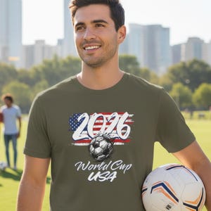 Puede incluir: Una camiseta verde oliva con el texto "2026 World Cup USA" y un gráfico de balón de fútbol. El diseño incorpora la bandera estadounidense. Una persona sostiene un balón de fútbol.