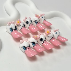 May include: A set of ten white and pink duck-themed artificial nails. Each nail features a duck face design with blue eyes, a pink beak, and a small pink bow or flower. The nails are arranged on a white surface.