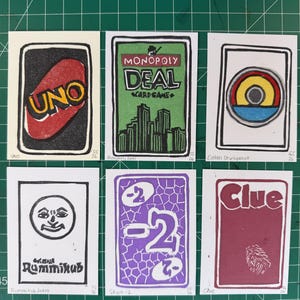 May include: Six rectangular art prints featuring hand-drawn illustrations of board and card game elements. The designs include Uno, Monopoly Deal, Catan Development, Rummikub Joker, Skyjo -2, and Clue, each labeled with its respective game title.