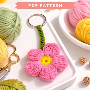 Puede incluir: Un llavero de flores de ganchillo rosa y amarillo. El llavero está sujeto a un anillo de metal. La imagen también muestra ovillos de lana verdes y amarillos, tijeras y el texto "Crochet Flower Keychain PDF Pattern."