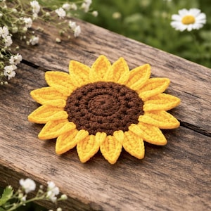 May include: A handmade crochet sunflower with yellow petals and a brown center. The sunflower is placed on a weathered wooden surface, with a blurred green background of foliage and white flowers.