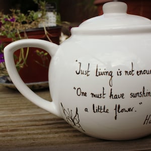 Puede incluir: Tetera de cerámica blanca con tapa y asa. La tetera tiene una cita escrita en escritura negra: "Solo vivir no es suficiente... Uno debe tener sol, libertad y una pequeña flor". Una mariposa está dibujada en el lateral.