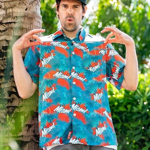 May include: A short-sleeved button-down shirt with a tropical design featuring the word "Miami" in white, red, and blue. The shirt has a white collar and is paired with a black cap.