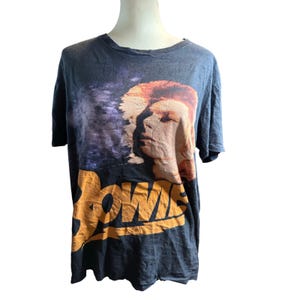 Könnte beinhalten: Dunkelgraues T-Shirt mit einem Porträt von David Bowie mit orangefarbenem Haar und dem Wort "BOWIE" in großen orangefarbenen Buchstaben. Das T-Shirt hat einen Rundhalsausschnitt und kurze Ärmel. Das T-Shirt wird auf einer Schaufenquin präsentiert.