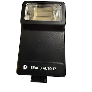 May include: Black Sears Auto 17 camera flash with a rectangular light and the text "SEARS AUTO 17". The flash has a rectangular light and a black body. The flash is designed for use with cameras.