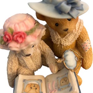May include: A figurine of two teddy bears, one wearing a pink hat with roses and the other a blue hat. They are looking at a photo album with bear photos. The bears are wearing pearl necklaces and lace collars.