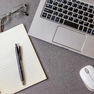 May include: A workspace scene with a silver laptop, a notebook with lined paper, a black pen, spectacles, and a white computer mouse. The items are arranged on a grey surface.
