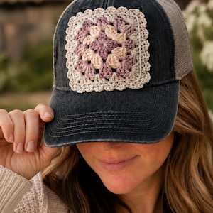 Può includere: Un cappellino da baseball nero antracite con una toppa quadrata all'uncinetto sul pannello frontale. La toppa presenta un motivo granny square multicolore nelle tonalità crema, viola chiaro e malva, rifinito con un bordo smerlato color crema.