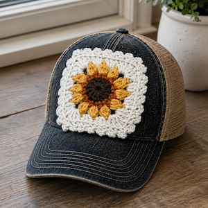 May include: A dark denim baseball cap featuring a square crochet patch on the front. The patch displays a sunflower design with a dark brown center, yellow petals, and a white border. The cap has a tan mesh back and a distressed denim brim.