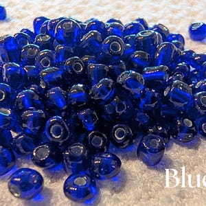 K&ouml;nnte beinhalten: Ein Haufen gl&auml;nzender, tiefblauer Glasperlen. Die Perlen sind rund mit einem Loch in der Mitte. Das Wort "Blue" ist in Wei&szlig; am unteren Rand des Bildes geschrieben. Diese Perlen eignen sich f&uuml;r die Schmuckherstellung und Bastelarbeiten.