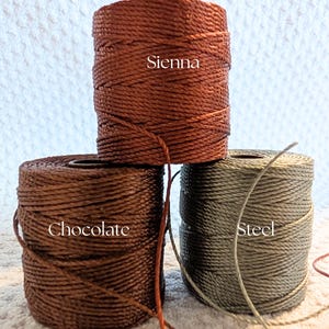 K&ouml;nnte beinhalten: Drei Spulen gewachster Kordel in den Farben Sienna, Schokolade und Stahl. Die Sienna-Spule ist rotbraun, die Schokoladen-Spule ist dunkelbraun und die Stahl-Spule ist ein ged&auml;mpftes Graugr&uuml;n. Die Farbbezeichnungen sind auf den Spulen aufgedruckt.