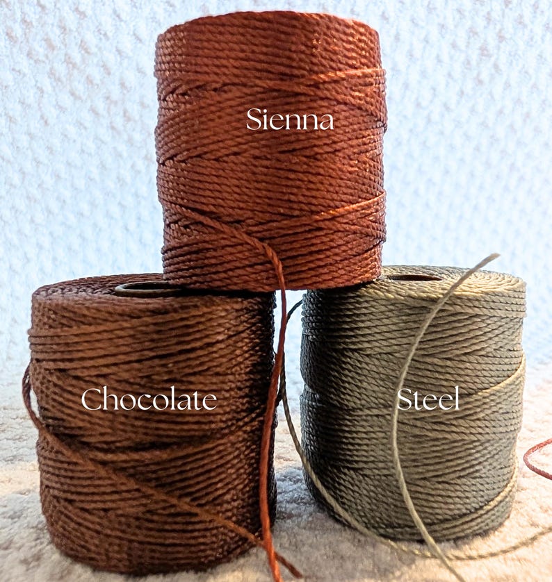 K&ouml;nnte beinhalten: Drei Spulen gewachster Kordel in den Farben Sienna, Schokolade und Stahl. Die Sienna-Spule ist rotbraun, die Schokoladen-Spule ist dunkelbraun und die Stahl-Spule ist ein ged&auml;mpftes Graugr&uuml;n. Die Farbbezeichnungen sind auf den Spulen aufgedruckt.