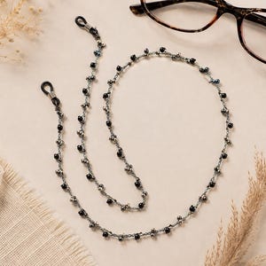 K&ouml;nnte beinhalten: Eine Perlen-Brillenkette mit schwarzen und silbernen Perlen. Die Kette liegt in der N&auml;he einer Brille mit Schildpattmuster. Die Kette hat Schlaufen an jedem Ende, um sie an der Brille zu befestigen. Die Kette ist etwa 71 cm lang.