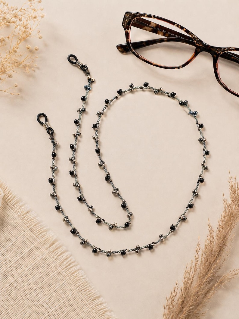 K&ouml;nnte beinhalten: Eine Perlen-Brillenkette mit schwarzen und silbernen Perlen. Die Kette liegt in der N&auml;he einer Brille mit Schildpattmuster. Die Kette hat Schlaufen an jedem Ende, um sie an der Brille zu befestigen. Die Kette ist etwa 71 cm lang.