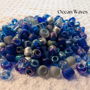 K&ouml;nnte beinhalten: Eine Ansammlung von Glasperlen in Blau-, Silber- und T&uuml;rkist&ouml;nen. Die Perlen variieren in Form und Gr&ouml;&szlig;e, einige haben ein mattiertes Aussehen. Der Text "Ocean Waves" ist &uuml;ber den Perlen zu sehen.