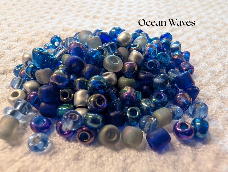 K&ouml;nnte beinhalten: Eine Ansammlung von Glasperlen in Blau-, Silber- und T&uuml;rkist&ouml;nen. Die Perlen variieren in Form und Gr&ouml;&szlig;e, einige haben ein mattiertes Aussehen. Der Text "Ocean Waves" ist &uuml;ber den Perlen zu sehen.