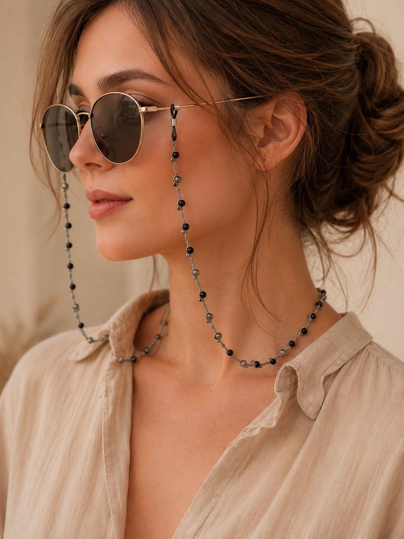 K&ouml;nnte beinhalten: Runde Sonnenbrille mit goldenem Rahmen und dunklen Gl&auml;sern, kombiniert mit einer Perlen-Brillenkette. Die Kette besteht aus abwechselnd schwarzen und grauen Perlen und bietet ein modisches Accessoire f&uuml;r die Brille.