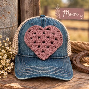 May include: A blue denim baseball cap featuring a large, mauve-colored crocheted heart appliqué centered on the front panel. The cap includes a mesh back and a curved brim with visible stitching. A label above the cap displays the text Mauve.