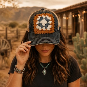 May include: A distressed dark blue denim baseball cap featuring a tan mesh back and a square crochet patch on the front. The patch is composed of a white center, a dark blue middle layer, and a rust-colored outer border in a classic granny square pattern.