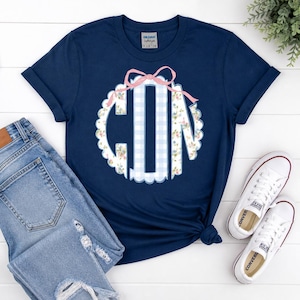 May include: Navy blue t-shirt with a monogram design. The monogram is a scalloped circle with a light blue gingham pattern and floral accents. A pink bow sits atop the monogram. Light blue jeans and white Converse sneakers are also in the image.