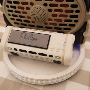 May include: A white and gray portable speaker with the name "Phillips" printed on the front. The speaker sits on a white decorative tray, with a larger tan speaker in the background.