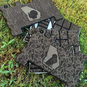 May include: A stack of flat, irregularly shaped decorative panels featuring a brown and black camouflage pattern. Each panel displays a light-colored outline of the state of Georgia with a black silhouette of a turkey or a dog inside the shape.