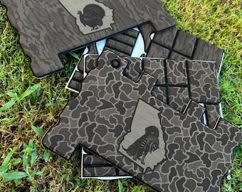 May include: A stack of flat, irregularly shaped decorative panels featuring a brown and black camouflage pattern. Each panel displays a light-colored outline of the state of Georgia with a black silhouette of a turkey or a dog inside the shape.