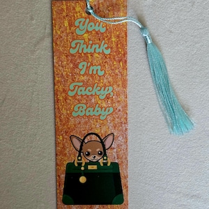 May include: A rectangular bookmark with a textured orange and gold background featuring the text You Think I'm Tacky, Baby in light green letters. Below the text is an illustration of a small brown dog inside a dark green handbag with a light blue tassel.