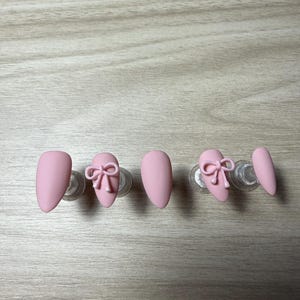 K&ouml;nnte beinhalten: Rosa mandelf&ouml;rmige Press-On-N&auml;gel. Zwei N&auml;gel weisen ein 3D-Schleifendesign auf. Die N&auml;gel werden auf transparenten Kunststoffst&auml;ndern vor einer hellen Holzmaserung pr&auml;sentiert. Die N&auml;gel haben ein mattes Finish.