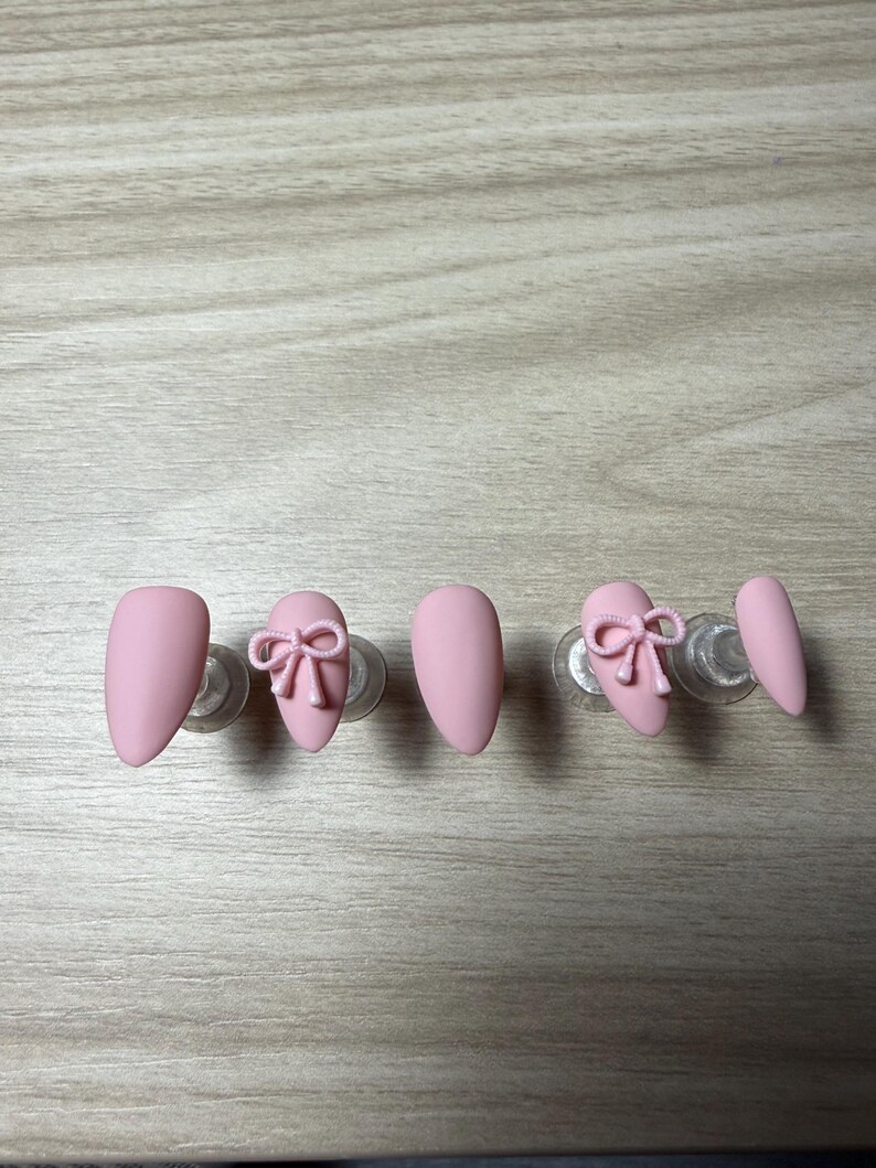 K&ouml;nnte beinhalten: Rosa mandelf&ouml;rmige Press-On-N&auml;gel. Zwei N&auml;gel weisen ein 3D-Schleifendesign auf. Die N&auml;gel werden auf transparenten Kunststoffst&auml;ndern vor einer hellen Holzmaserung pr&auml;sentiert. Die N&auml;gel haben ein mattes Finish.