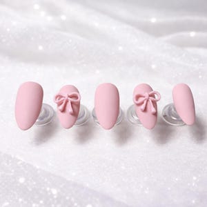 K&ouml;nnte beinhalten: F&uuml;nf rosa Kunstn&auml;gel, jeweils auf einem transparenten Kunststoffst&auml;nder befestigt. Zwei N&auml;gel weisen ein 3D-Schleifendesign auf. Die N&auml;gel haben eine ovale Form und ein mattes Finish, vor einem wei&szlig;en, glitzernden Hintergrund.