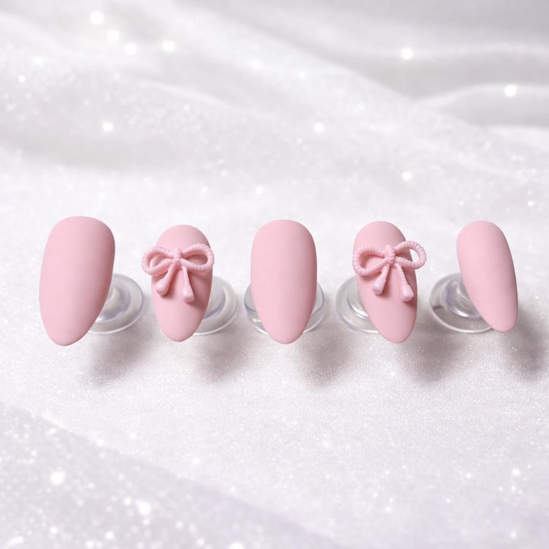 K&ouml;nnte beinhalten: F&uuml;nf rosa Kunstn&auml;gel, jeweils auf einem transparenten Kunststoffst&auml;nder befestigt. Zwei N&auml;gel weisen ein 3D-Schleifendesign auf. Die N&auml;gel haben eine ovale Form und ein mattes Finish, vor einem wei&szlig;en, glitzernden Hintergrund.