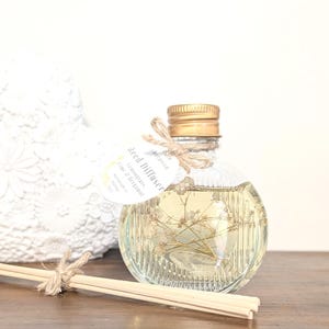 May include: A round, ribbed glass reed diffuser bottle filled with light yellow liquid and dried flowers. It features a gold screw cap, a jute twine bow, and a white circular tag labelled "Pure Botanica Reed Diffuser Lemongrass, Lime & Bergamot".