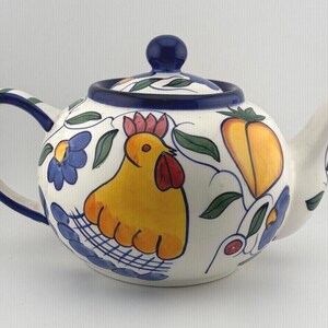 Puede incluir: Una tetera de cerámica con base color crema que presenta un diseño pintado a mano de un gallo amarillo con cresta roja, flores azules y frutas amarillas. La tetera tiene un asa azul oscuro, una tapa a juego con pomo y un pico curvo.