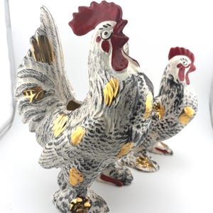 Peut inclure: Deux figurines de coqs en céramique avec une base blanche, des détails mouchetés noirs, une crête et des barbillons rouge foncé, ainsi que des accents dorés métalliques sur les plumes et les pattes. Les coqs sont alignés vers la droite.