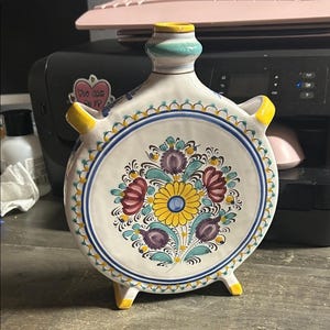 Puede incluir: Una botella de cerámica con un diseño floral. La botella es blanca con un cuerpo redondo y un cuello estrecho. El diseño floral presenta flores amarillas, rojas, moradas y azules. La botella tiene asas y pies amarillos.
