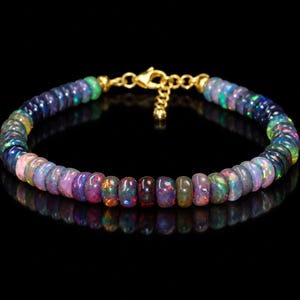 Op de afbeelding: Een armband bestaande uit kleine, ronde, donkergekleurde opaal kralen met iriserende flitsen van groen, blauw, rood en paars. Het sieraad is voorzien van een goudkleurige metalen karabijnsluiting en een verstelbare kettingverlenging.
