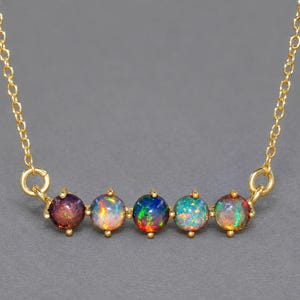 Op de afbeelding: Een gouden ketting met een horizontale balk van vijf ronde, in klauwen gezette opalen. Elke opaal vertoont een uniek kleurenspel, inclusief flitsen van rood, groen, blauw en oranje, hangend aan een fijne gouden schakelketting met ronde verbindingsstukken.