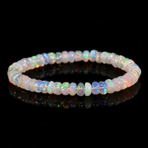 Puede incluir: Una pulsera circular compuesta por pequeñas cuentas de ópalo translúcidas en forma de disco. Cada cuenta muestra un juego de colores con destellos iridiscentes verdes, azules y rosas sobre una base blanca lechosa, ensartadas en un bucle.
