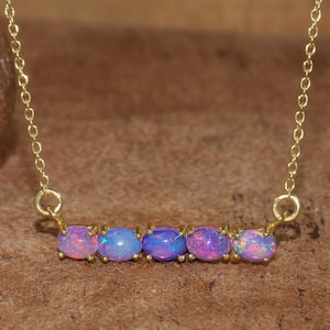 May include: A delicate gold necklace featuring five oval opals in shades of pink and blue. The opals are set in individual gold prongs and are connected by a fine gold chain. The necklace is displayed on a brown surface.