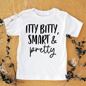 May include: A white t-shirt with the words "ITTY BITTY, SMART & pretty" in black script. The shirt is displayed on a mustard-coloured surface, with dried plant stems and leaves arranged around it.
