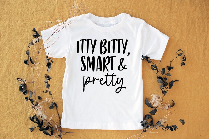 May include: A white t-shirt with the words "ITTY BITTY, SMART & pretty" in black script. The shirt is displayed on a mustard-coloured surface, with dried plant stems and leaves arranged around it.