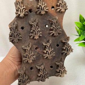 May include: A rectangular brown wooden block with a carved relief pattern featuring twelve repeating floral motifs arranged in a grid. The surface of the wood shows natural grain and texture, with small circular holes interspersed between the carvings.