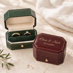 May include: Two octagonal velvet ring boxes, one dark green and one deep burgundy. The green box is open, revealing a gold band and a diamond ring. The burgundy box is closed, featuring gold text reading Olivia & Noah and 09.14.2026.