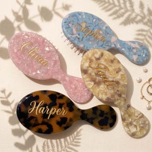 May include: Four oval hairbrushes with handles, each featuring a unique pattern and a gold-colored name printed in script. The brushes include a pink marble pattern labeled Olivia, a blue marble pattern labeled Sophia, a beige marble pattern labeled Jade, and a brown tortoiseshell pattern labeled Harper.