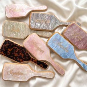 Könnte beinhalten: Eine Sammlung rechteckiger Haarbürsten mit Griffen, die verschiedene Marmormuster in Rosa, Blau, Grün und braunem Schildpatt aufweisen. Jede Bürste ist mit goldfarbenen Namen in Schreib- oder Blockschrift personalisiert, darunter Olivia, Sarah, Isla, Lily, Harper, Amelia und Sophia.
